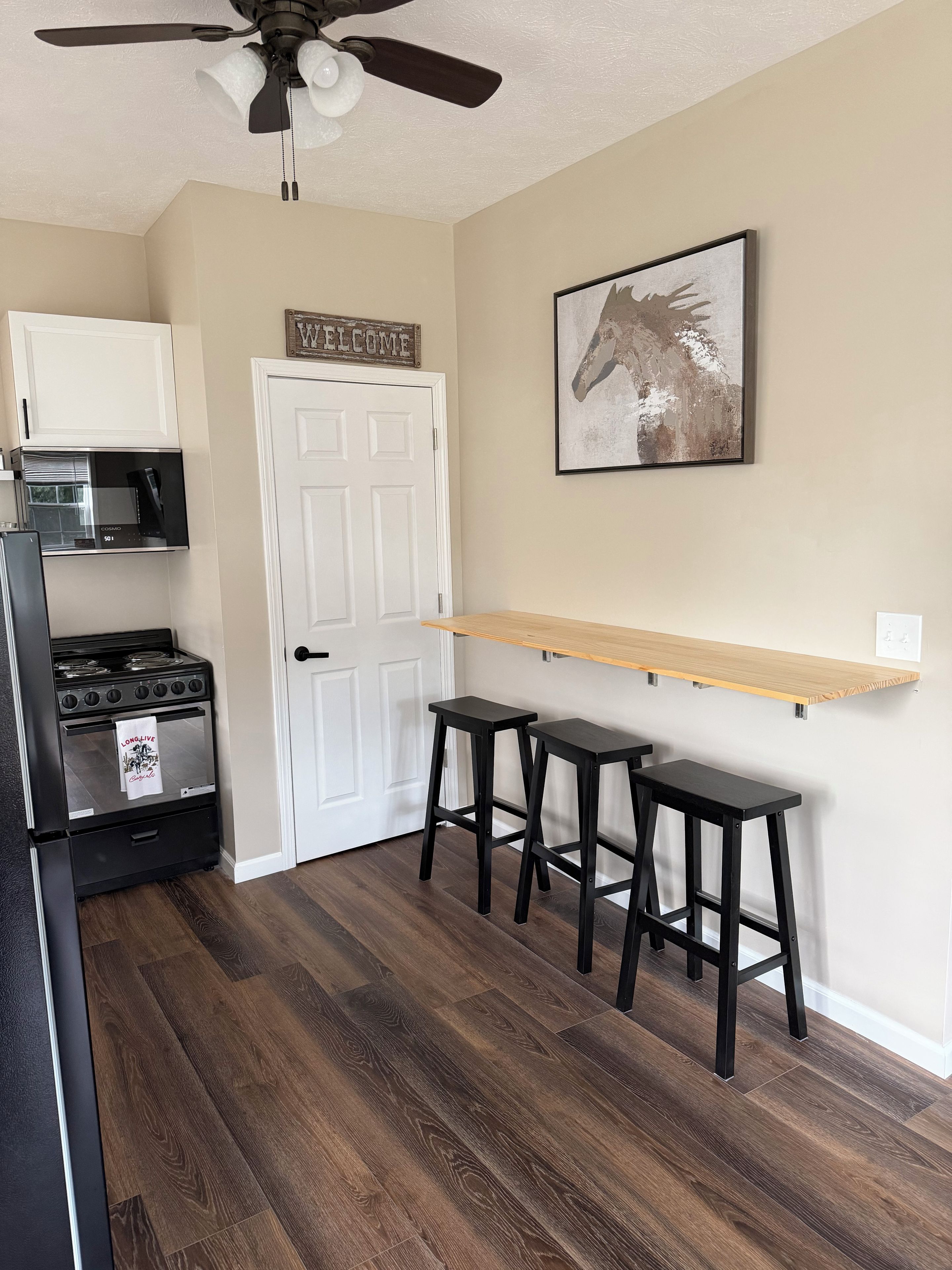 Una cocina con refrigerador negro, estufa y microondas, taburetes de madera, una puerta blanca y un cartel de bienvenida.