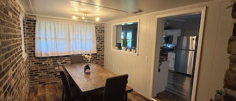 A dining area with a wooden table and chairs, a brick wall, and a view into the kitchen.