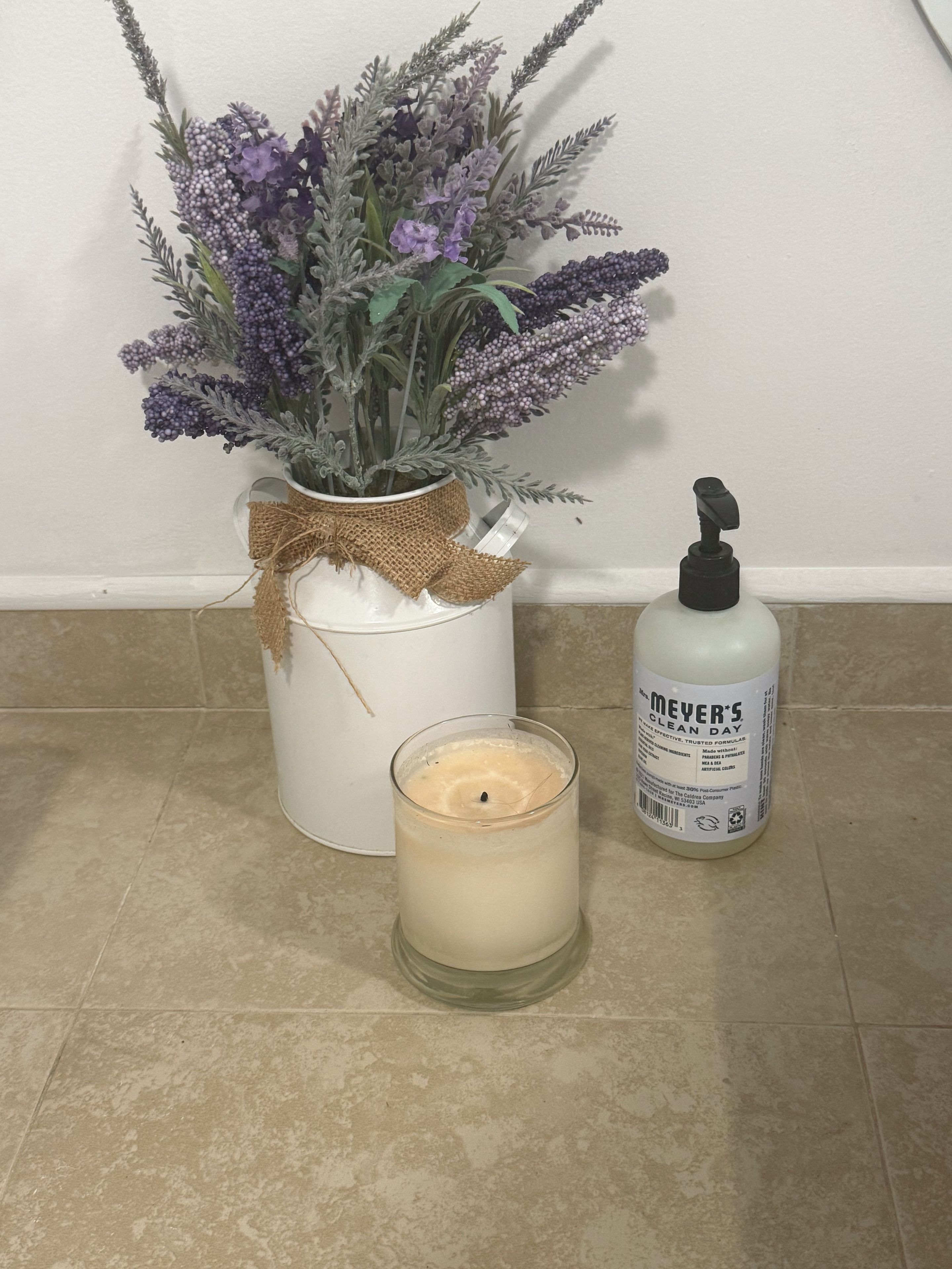 A white vase with a burlap bow, a glass candle, and a bottle of Meyer’s Clean Day on a tiled floor.