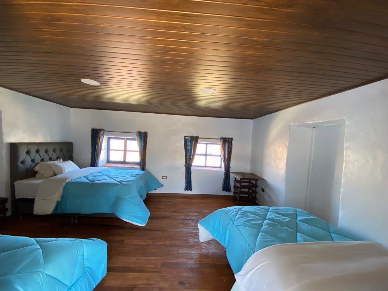 Una habitación con dos camas, piso de madera, techo con diseño de listones y ventana con cortinas.