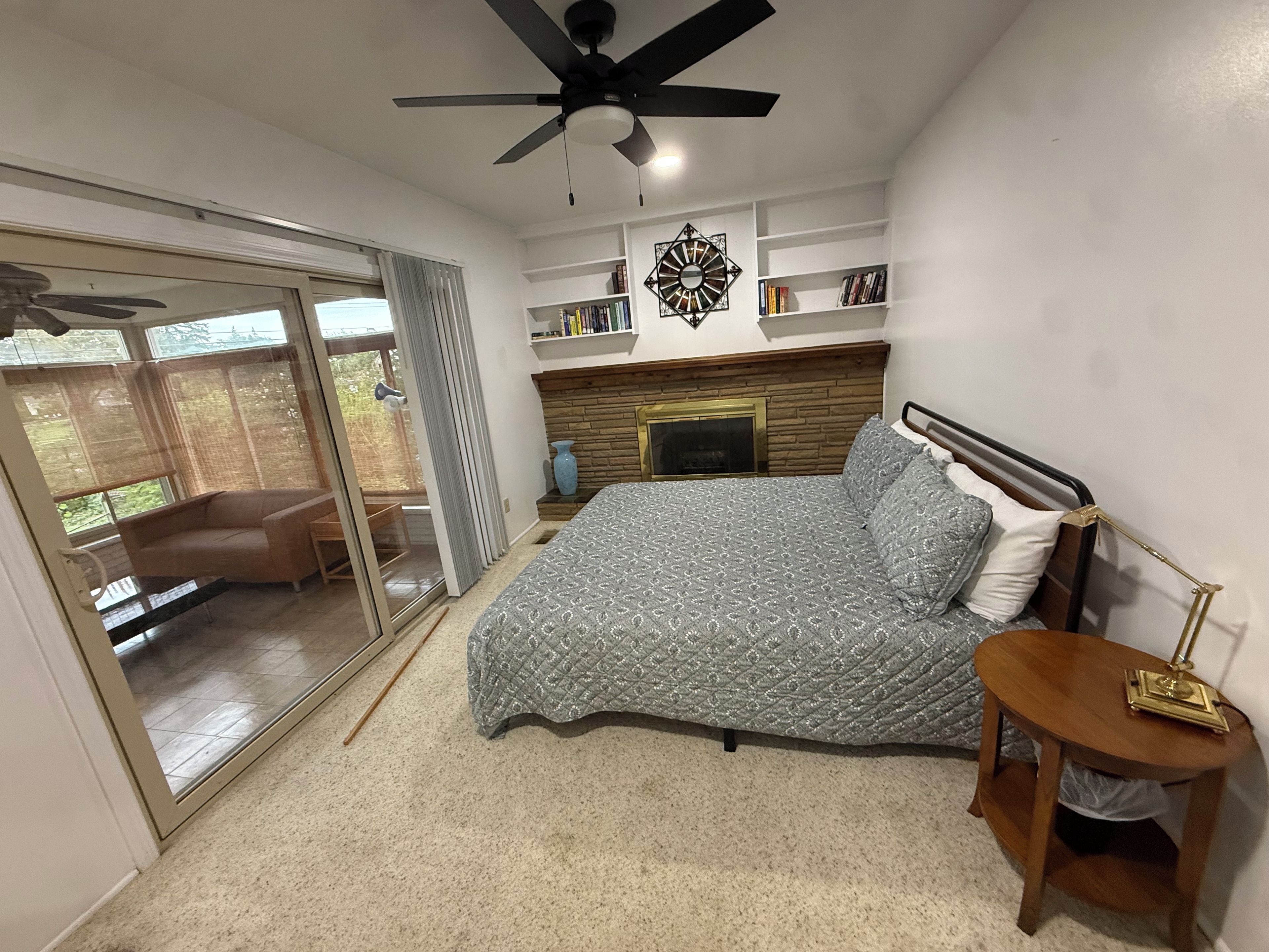 Une chambre à coucher comprenant un lit, une table de chevet, un ventilateur de plafond, une cheminée et une porte-fenêtre coulissante donnant sur un balcon.