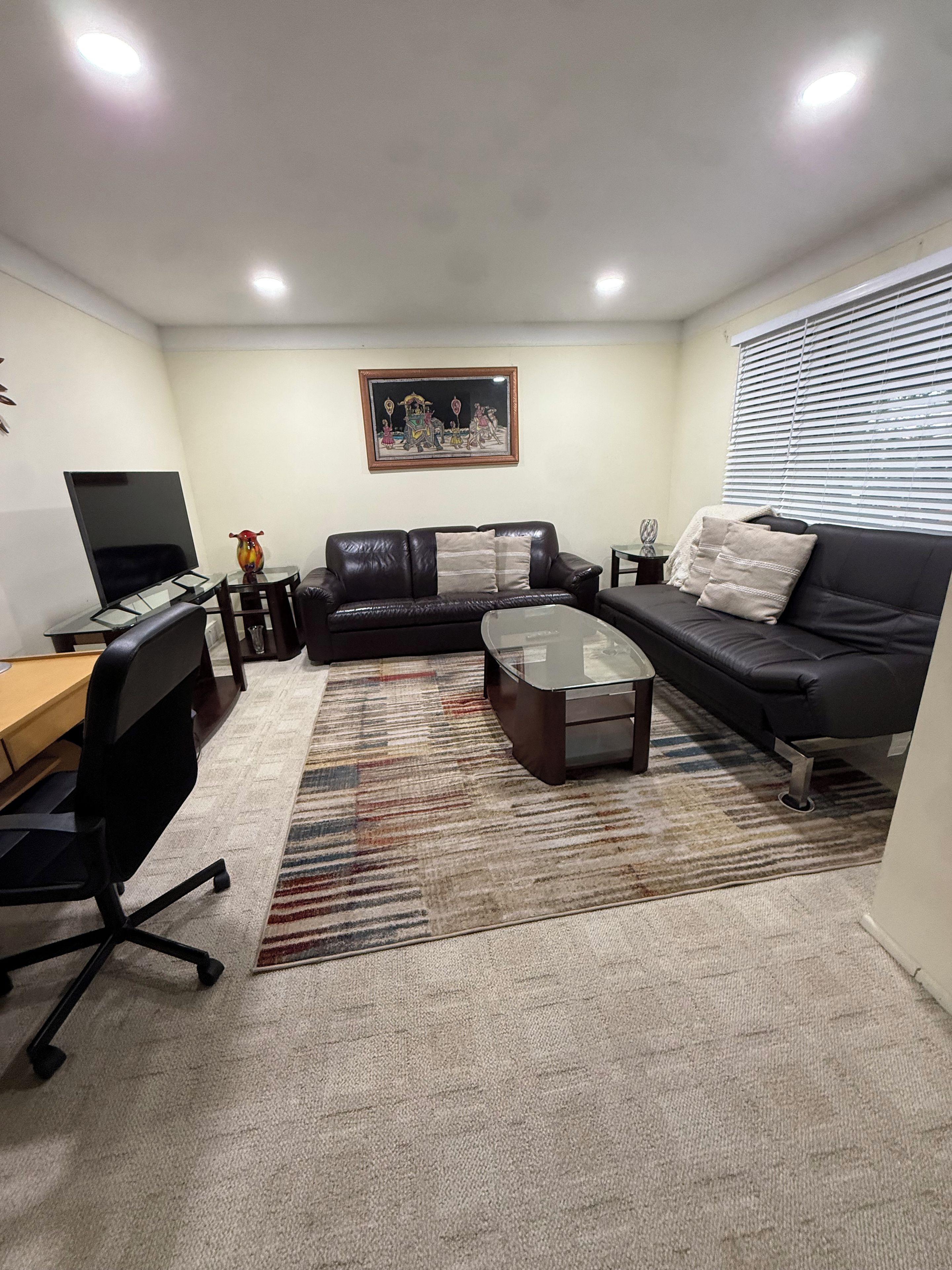 A living room with a black leather sofa, a glass coffee table, a black office chair, a TV, and a framed picture on the wall.