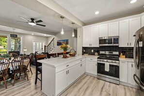 A modern kitchen with white cabinetry, stainless steel appliances, and a dining area with wooden chairs and a table. - Historic Borden House: Hot Tub Sauna Pet Friendly (Galveston)