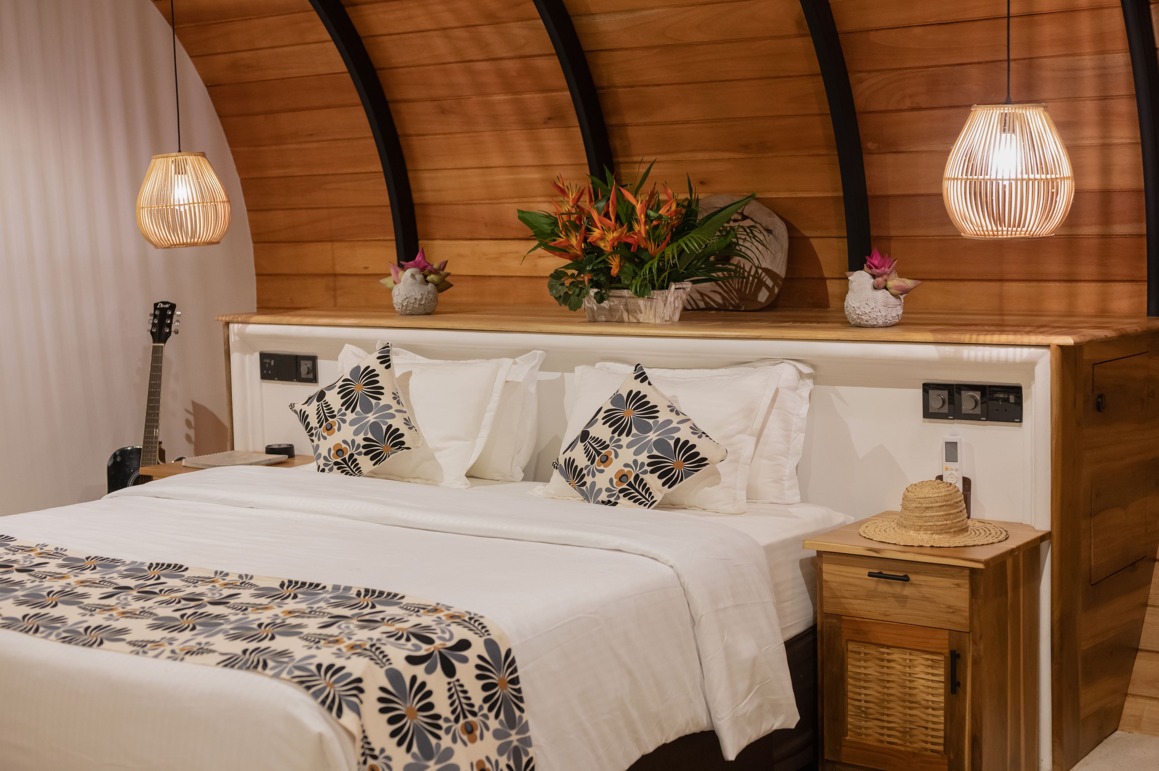 A bedroom with a wooden ceiling, a bed with white bedding, decorative pillows, a bedside table with a hat, and a guitar in the background.