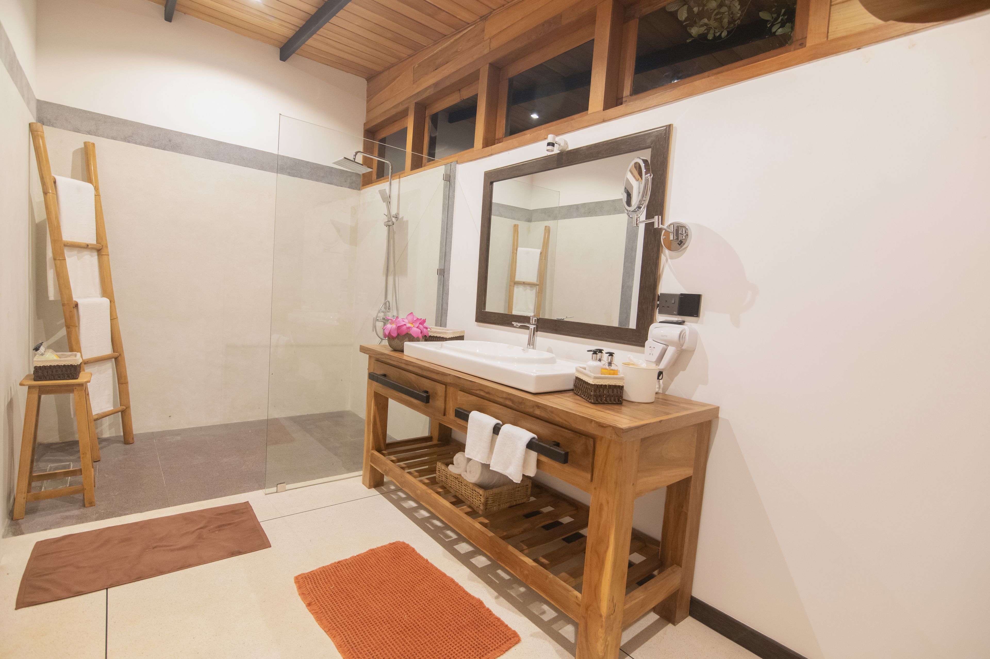 Ein Badezimmer mit einem Holzbadezimmerhocker, einem großen Spiegel, einer Dusche und einem Handtuchhalter.