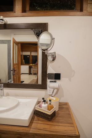 Un baño con un lavabo blanco, un espejo, un secador de pelo y artículos de aseo sobre un mueble de madera.
