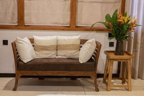 Un sofá marrón con cojines a rayas, una mesita de madera con un jarrón de flores y una ventana con persianas.