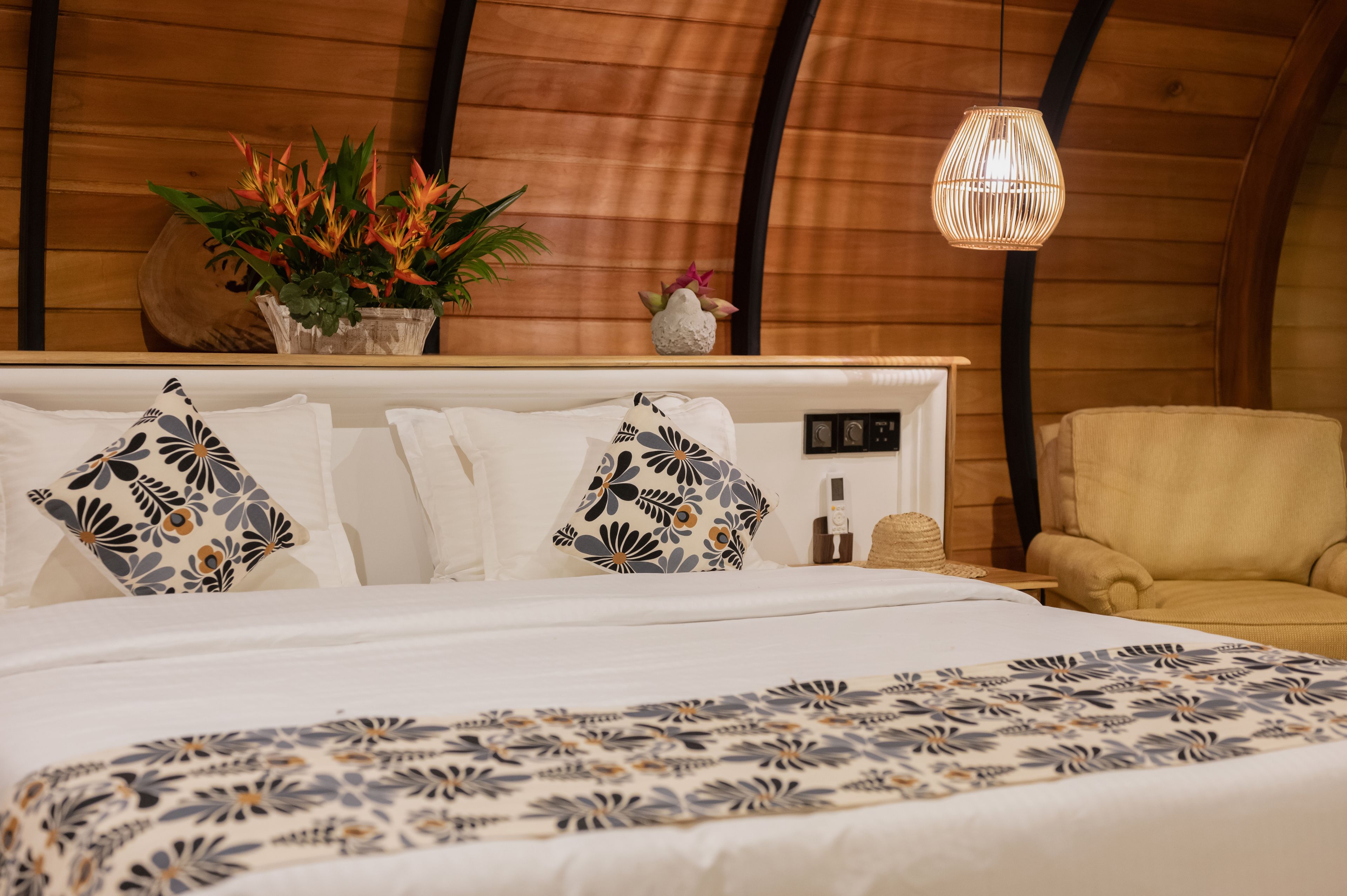 A bedroom with a wooden ceiling, a bed with patterned bedding, a bedside table with a lamp, and a chair.