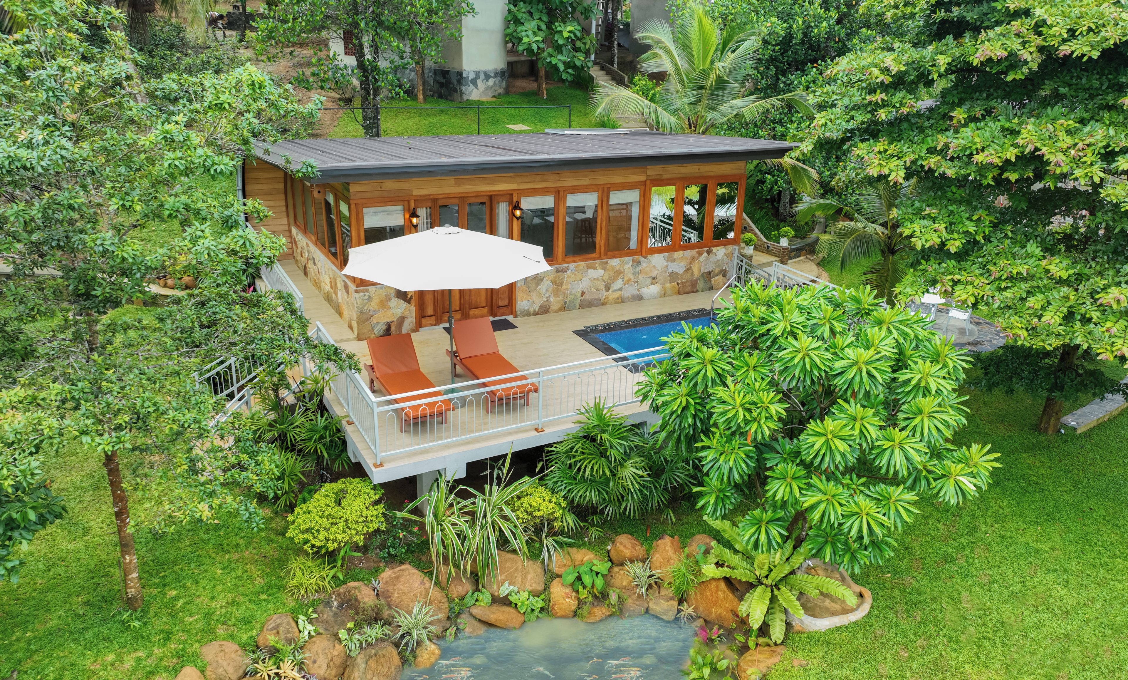 A two-story wooden house with a pool, surrounded by lush greenery and a small pond.