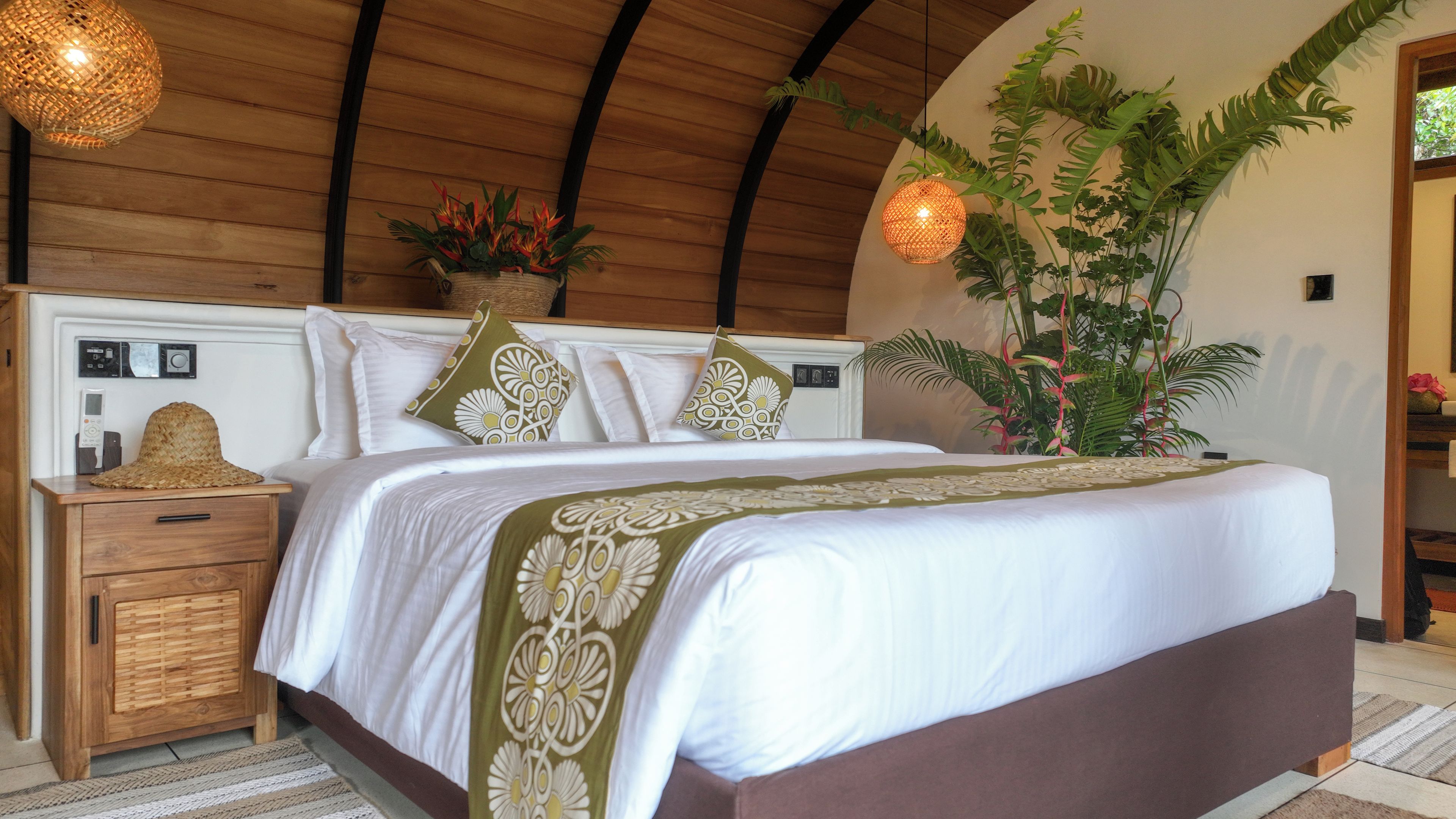 Un dormitorio con techo de madera, una cama con ropa de cama blanca y almohadas decorativas, una mesita de noche con un sombrero y un control remoto, y una planta grande.