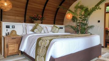 Un dormitorio con techo de madera, una cama con ropa de cama blanca y almohadas decorativas, una mesita de noche con un sombrero y un control remoto, y una planta grande.