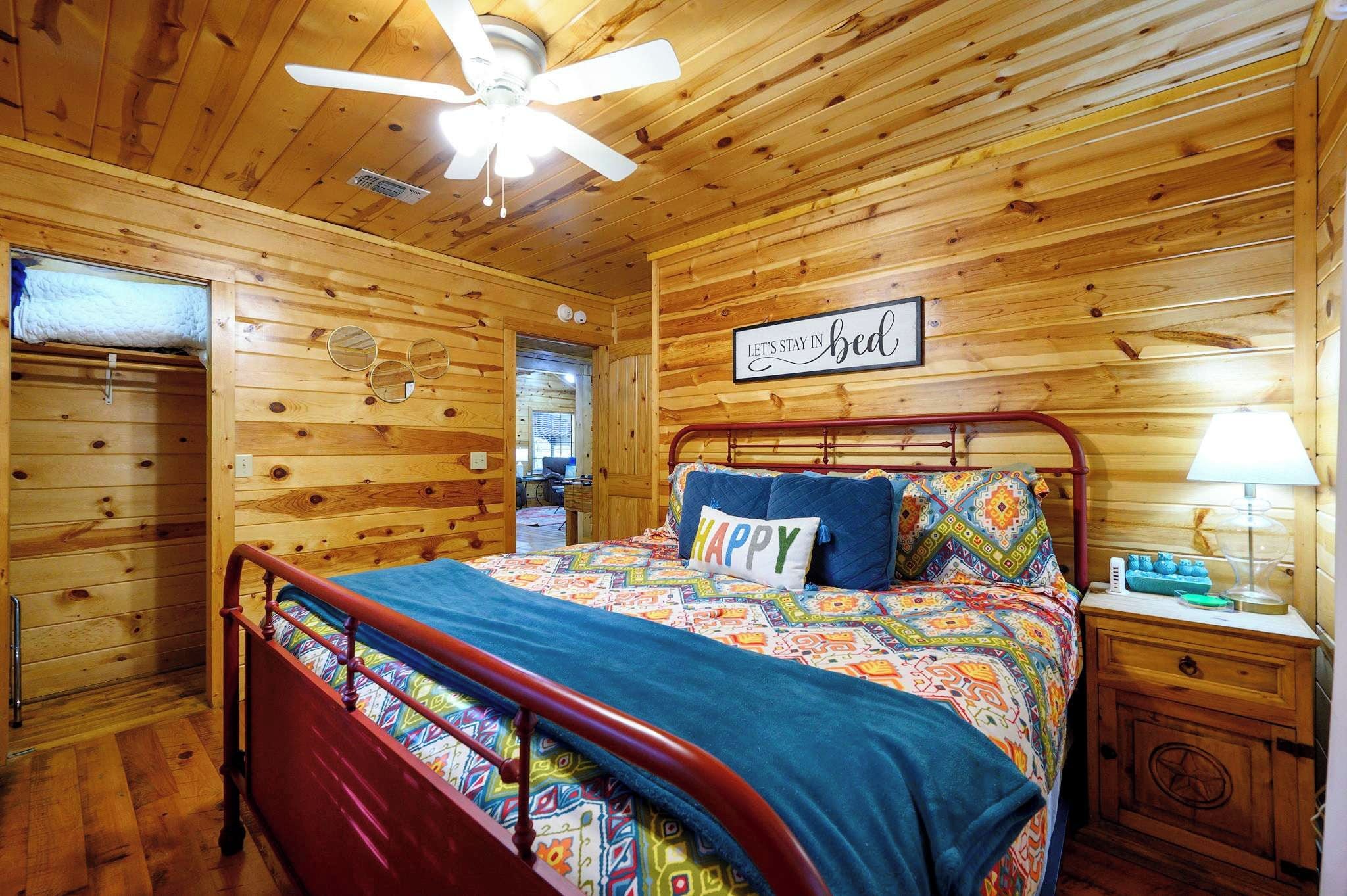 A wooden cabin bedroom with a bed, a nightstand, a ceiling fan, and a sign that says 'Let's stay in bed'.