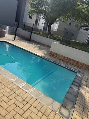 Una piscina con borde de azulejos, rodeada por un patio de ladrillo y una valla.