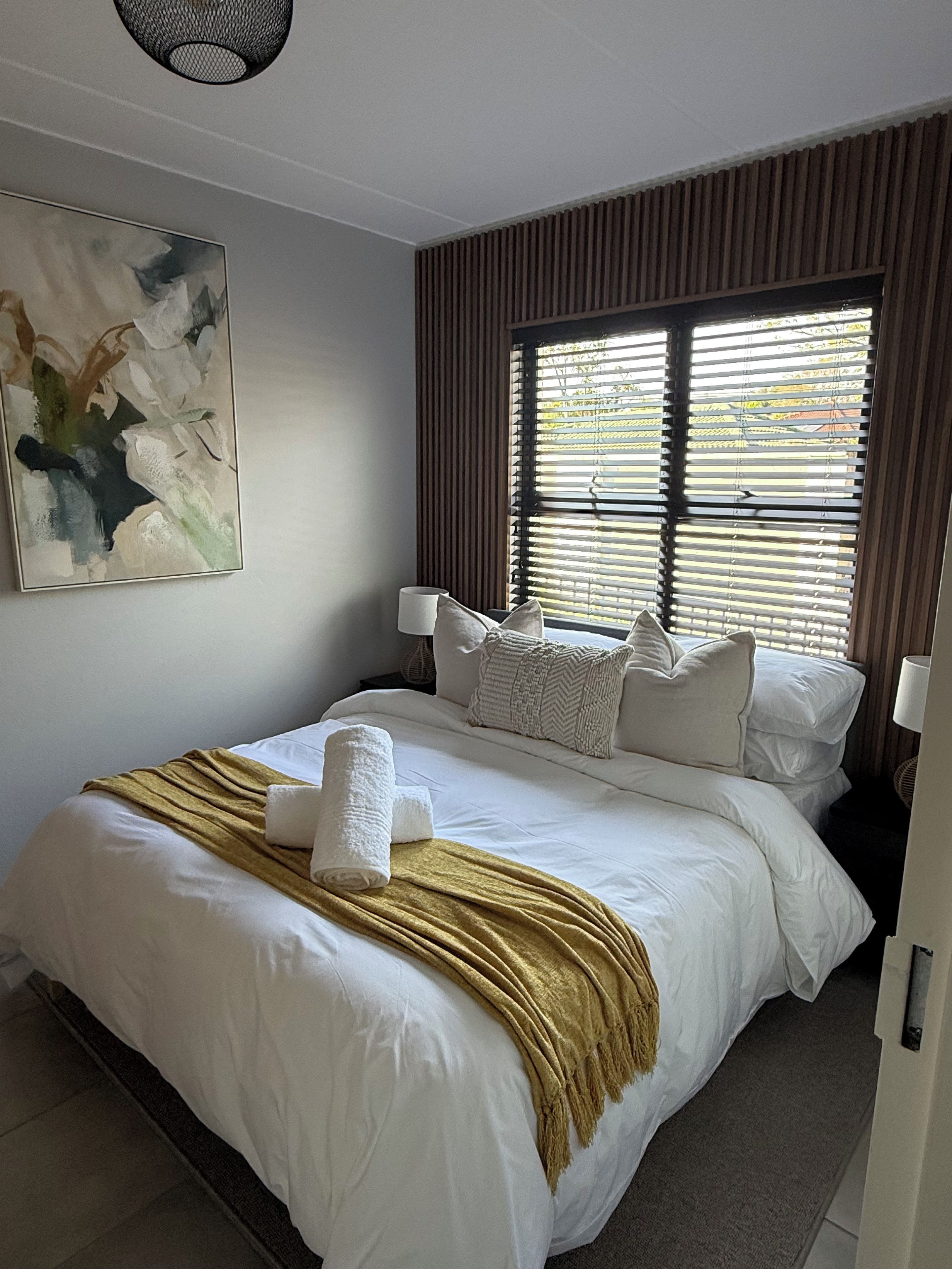 Ein Hotelzimmer mit einem großen Bett, einem Fenster mit Jalousien, einem Bild an der Wand und einer Nachttischlampe.