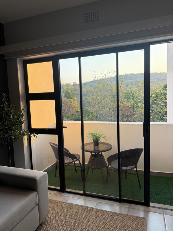 A room with a sliding glass door leading to a balcony with a table and chairs, a potted plant, and a small grassy area.