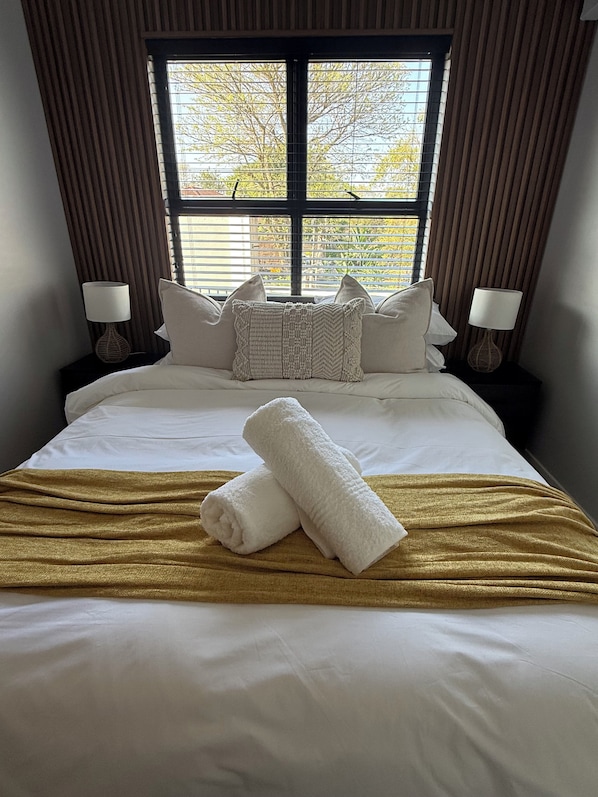 A neatly made bed with white linens and a gold throw blanket, flanked by two bedside lamps and a window with blinds.