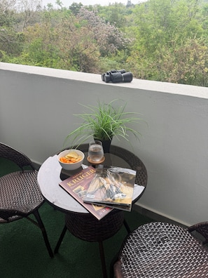 A balcony with a round table, two wicker chairs, a bowl of snacks, a glass of water, a potted plant, and a magazine.