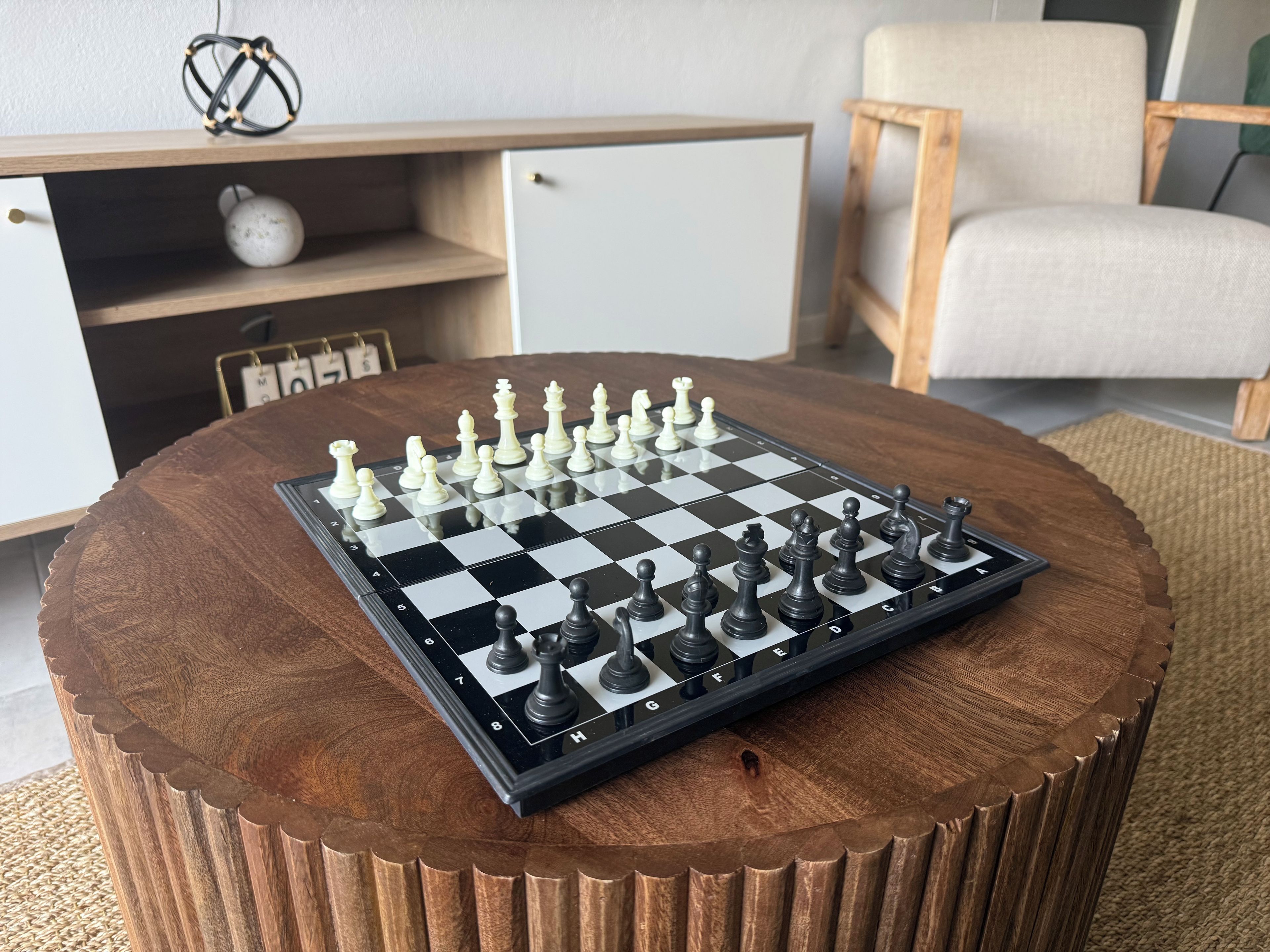 A chessboard on a round wooden table with a chair and a cabinet in the background.