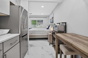 A compact kitchen with a refrigerator, a wooden dining table, and a bed with a window view.