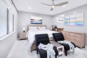 Un dormitorio moderno con una cama, mesitas de noche, un escritorio con sillas, un ventilador de techo y obras de arte en las paredes.