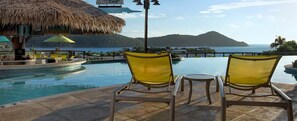 Zona de piscina con dos sillas amarillas y una sombrilla de paja, con vistas a una masa de agua y una isla distante.