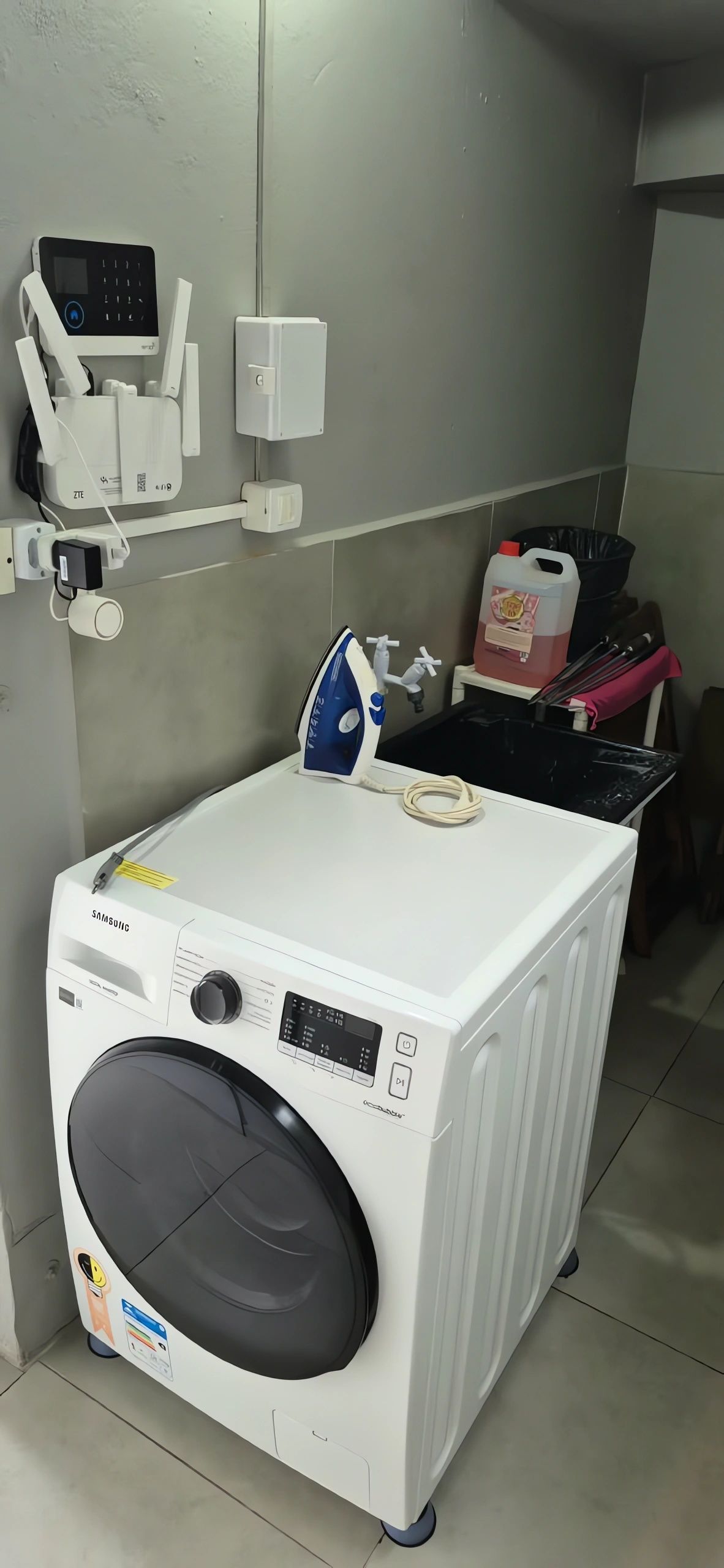 Eine kompakte, weiße Waschmaschine mit Frontlader, einem digitalen Display und Bedienfeld, ein an der Wand befestigtes Bügeleisen und eine kleine Ablage mit Reinigungsmitteln.