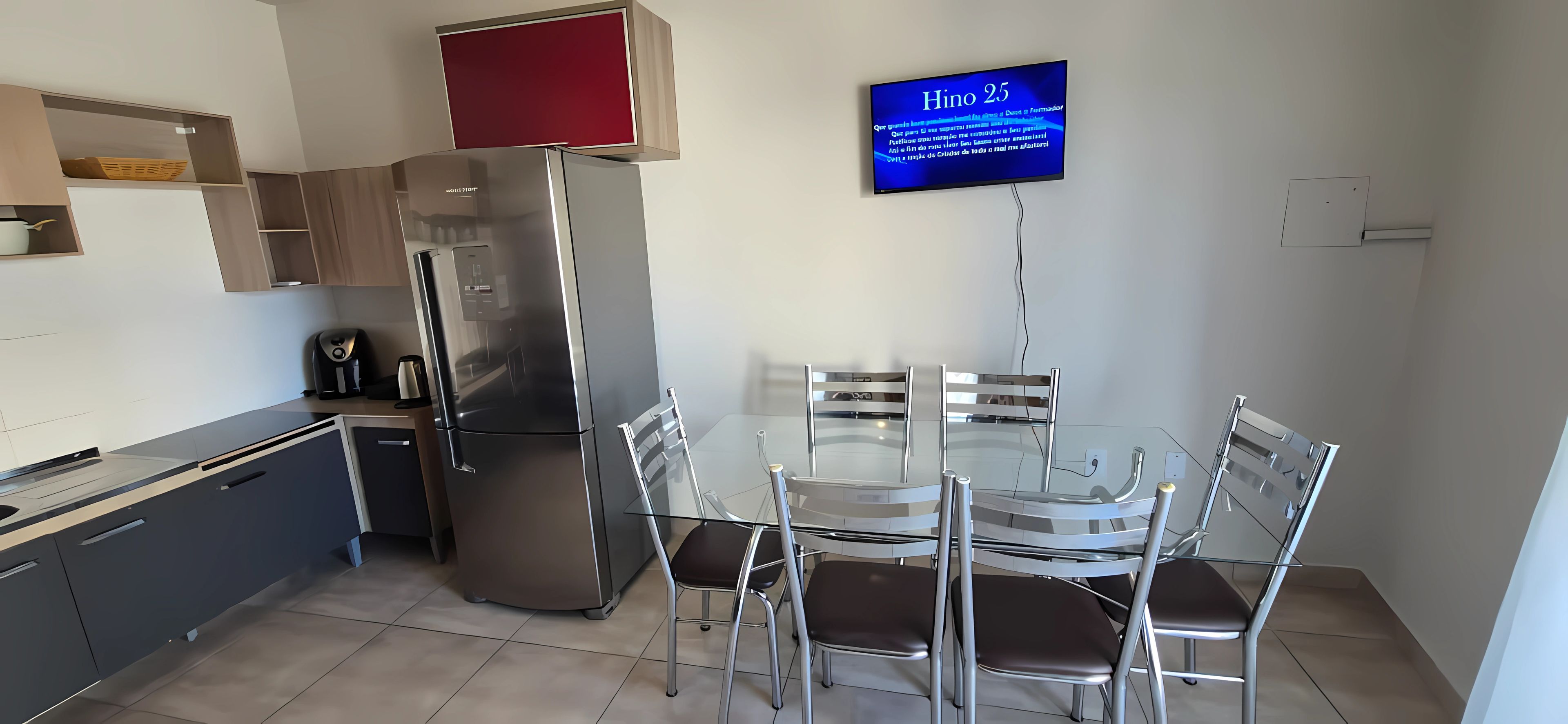 A modern kitchen with a stainless steel refrigerator, a glass dining table with chairs, and a flat-screen TV mounted on the wall.
