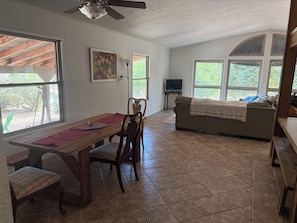 A dining area with a wooden table, chairs, and a red placemat. There is a television on a stand and a sofa with a blanket. - Riverside Horse Ranch Unplugged (Thornton)