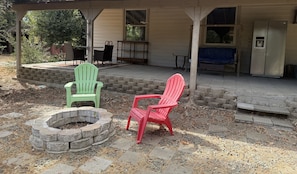A covered outdoor seating area with two Adirondack chairs, a stone fire pit, and a building with a window and a door.