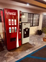 Máquina de Coca-Cola ao lado de um frigorífico, com uma janela e uma planta em vaso ao fundo.