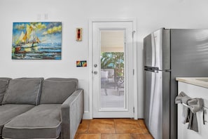 A compact living space with a grey sofa, a stainless steel refrigerator, a door with a window, and a painting of sailboats on the wall. - Siesta Villa | Private Patio | Dog Friendly (Port Aransas)