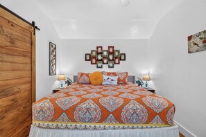 A bedroom with a large bed, two bedside lamps, a decorative wall piece, and a wooden door. - Siesta Villa | Private Patio | Dog Friendly (Port Aransas)