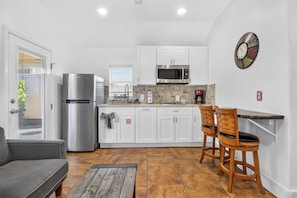 A compact kitchen with white cabinets, a stainless steel refrigerator, a microwave, and a coffee maker. There is a small dining area with two wooden chairs and a grey sofa. - Siesta Villa | Private Patio | Dog Friendly (Port Aransas)
