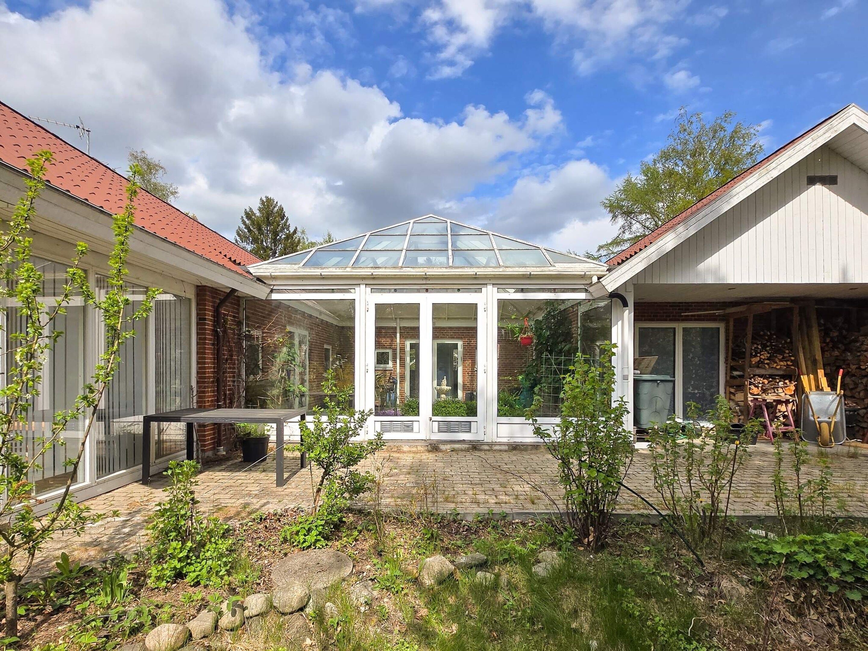 A covered patio with a glass roof, surrounded by a garden with various plants and a brick wall.