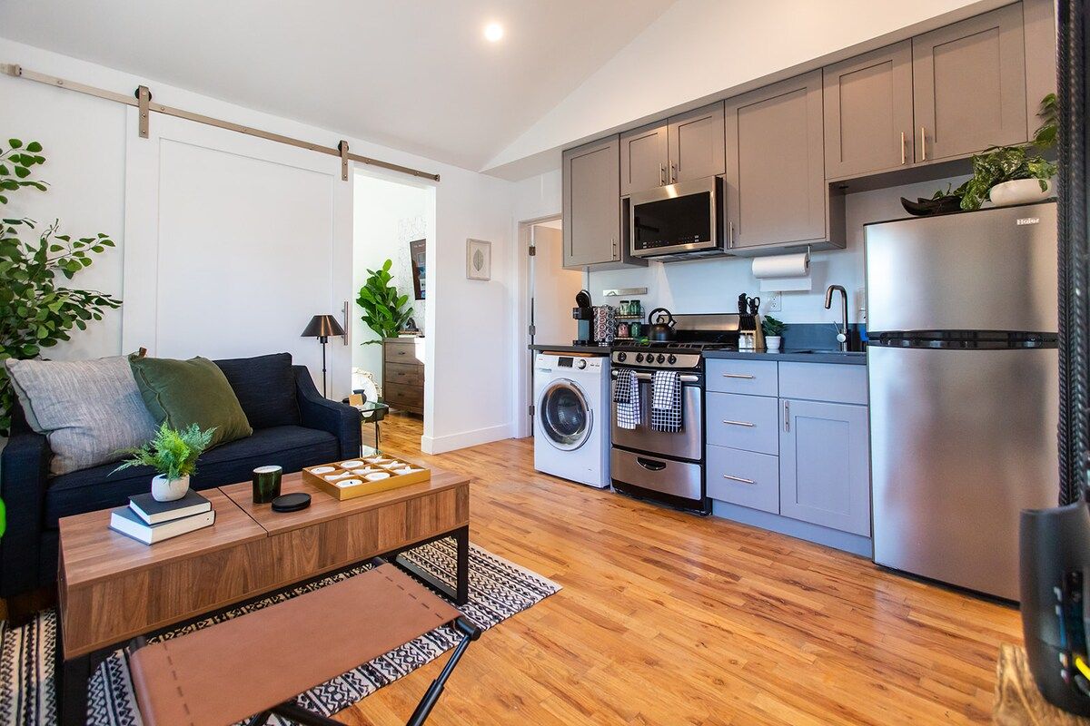 A compact living space with a sofa, coffee table, and kitchen area including a washing machine, oven, microwave, and refrigerator.