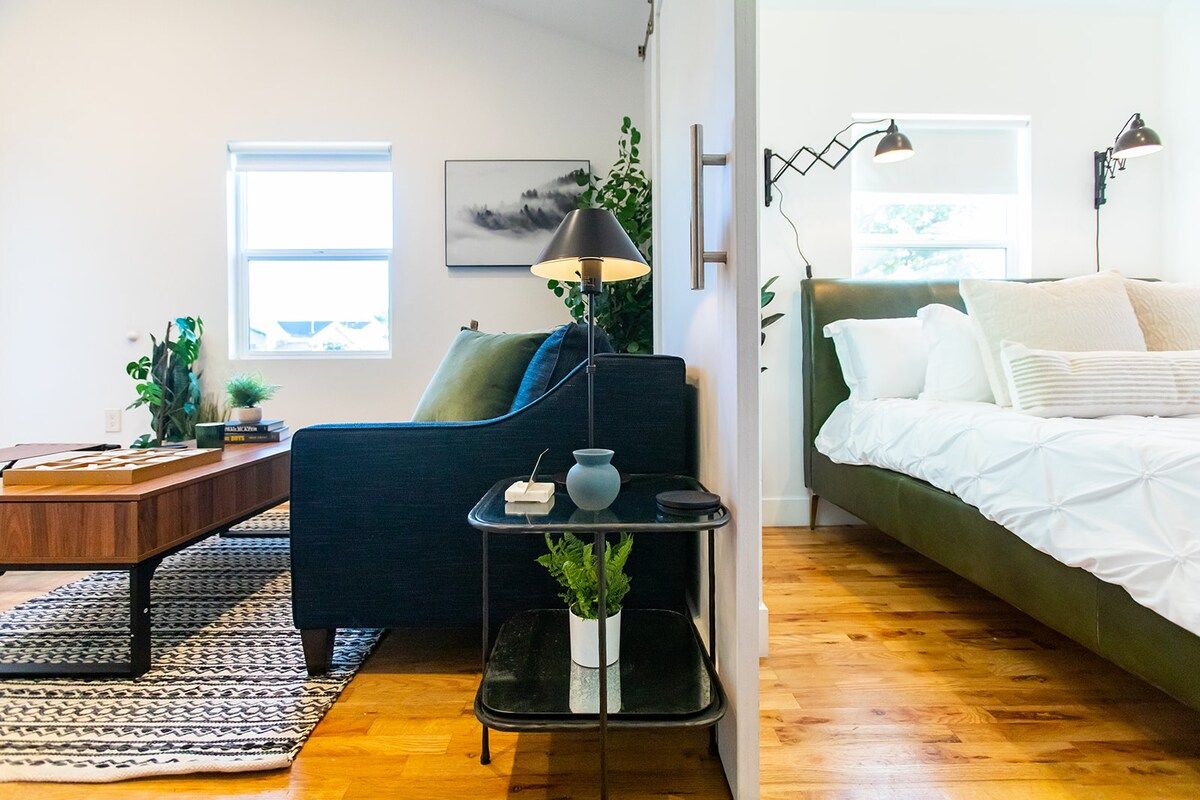 A modern bedroom with a green upholstered bed, a wooden desk, a black side table with a lamp, and a potted plant.