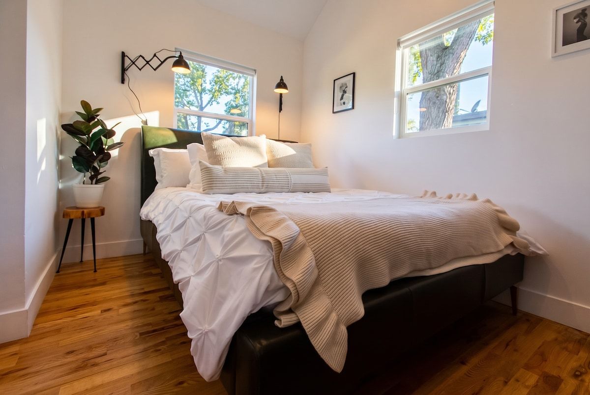 A bedroom with a bed, bedside tables, a potted plant, and two windows.
