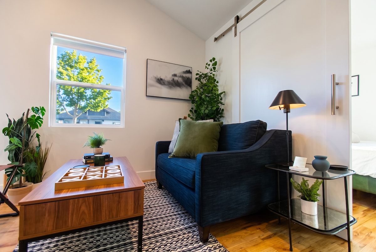 A modern living room with a blue sofa, a wooden coffee table, a floor lamp, and a view of a tree through a window.