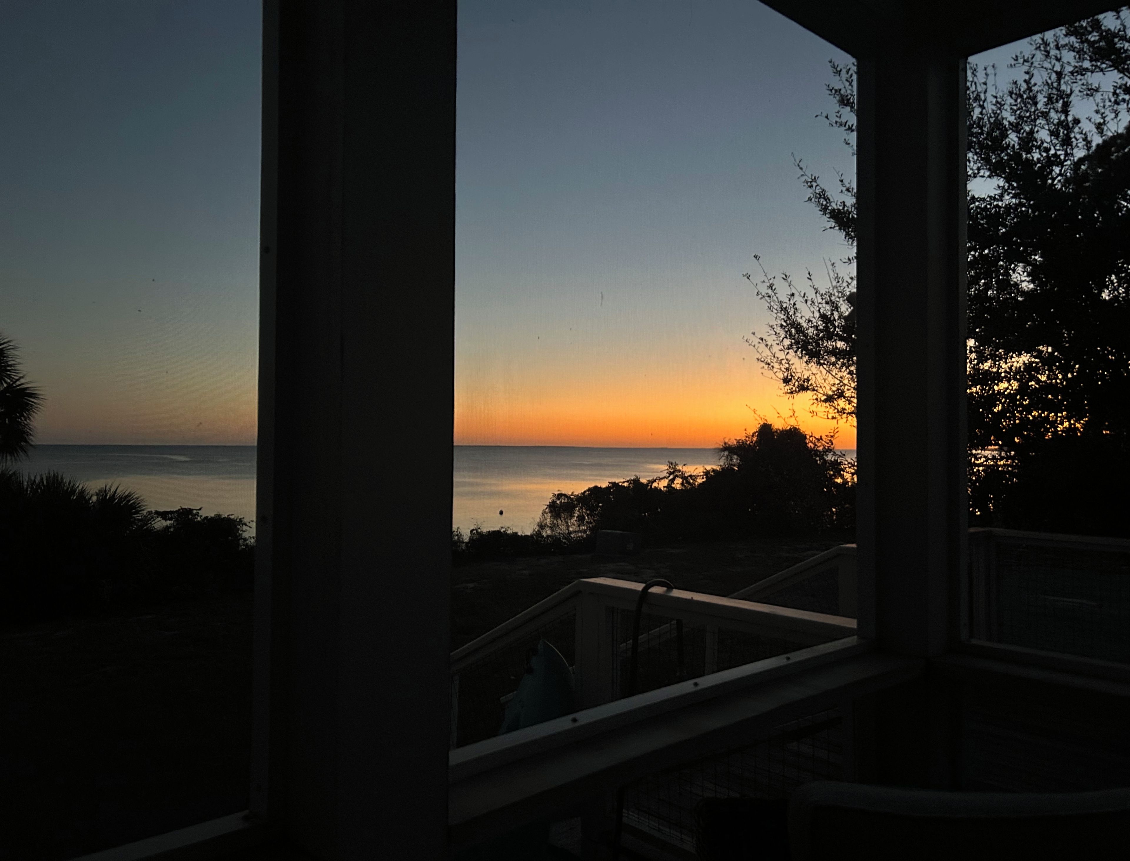 Näkymä ikkunasta, josta avautuu meren yllä auringonlaskun maisema. Etualalla on kaide ja kasvillisuutta.