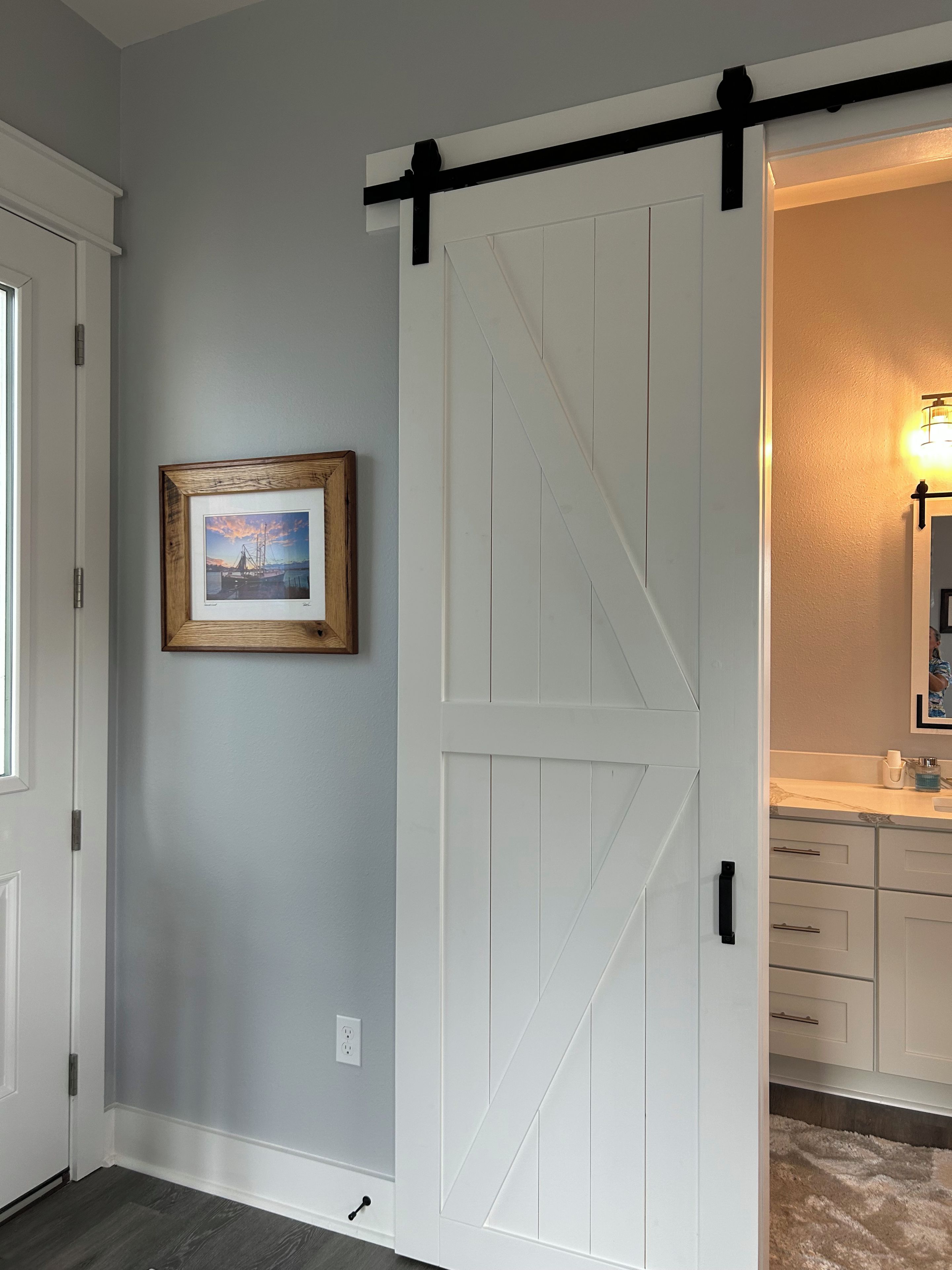A room with a white sliding barn door, a framed picture on the wall, and a bathroom with a sink and mirror.