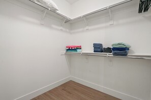 Un vestidor con paredes blancas y piso de madera. Contiene una barra metálica con ropa doblada y un estante blanco.