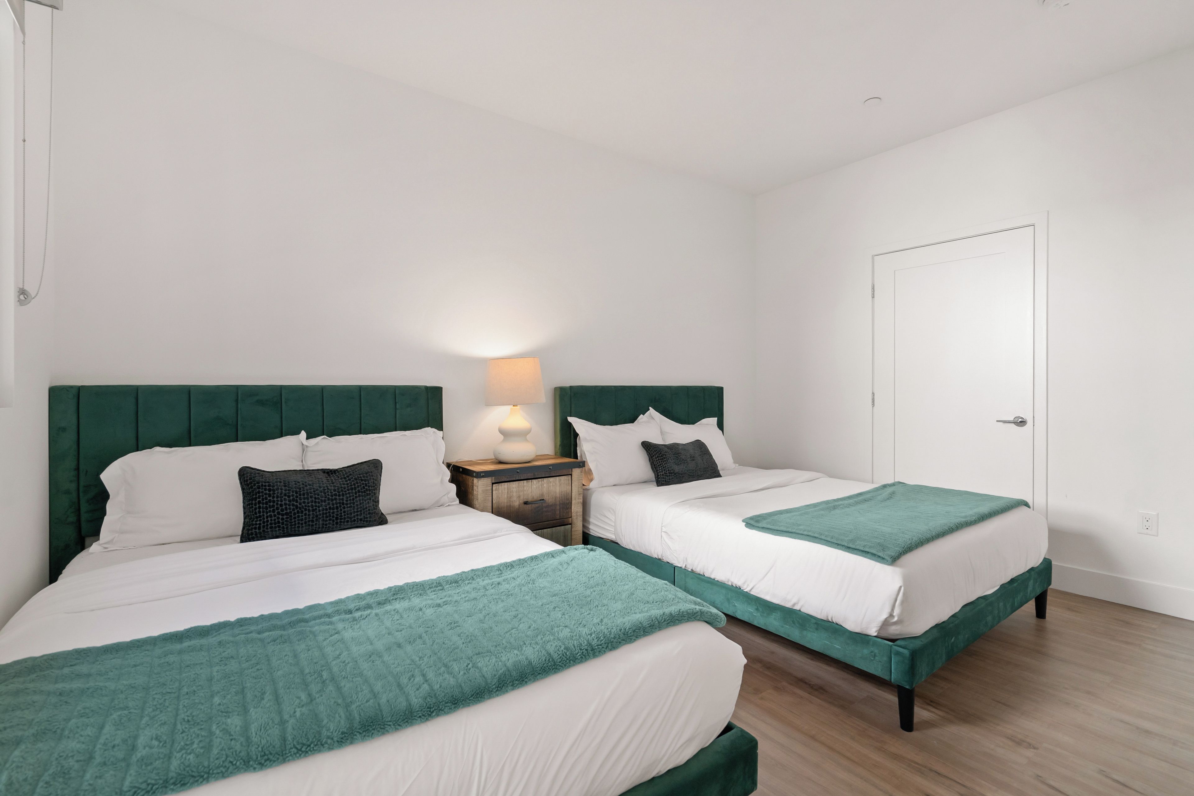 A bedroom with two beds, each with a green headboard, white bedding, and a teal throw blanket. There is a nightstand with a lamp between the beds.