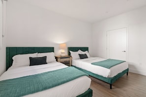 A bedroom with two beds, each with a green headboard, white bedding, and a teal throw blanket. There is a nightstand with a lamp between the beds. - The Amber Serene -  Peaceful 2BR Retreat - Pool & Gym (Santa Monica)