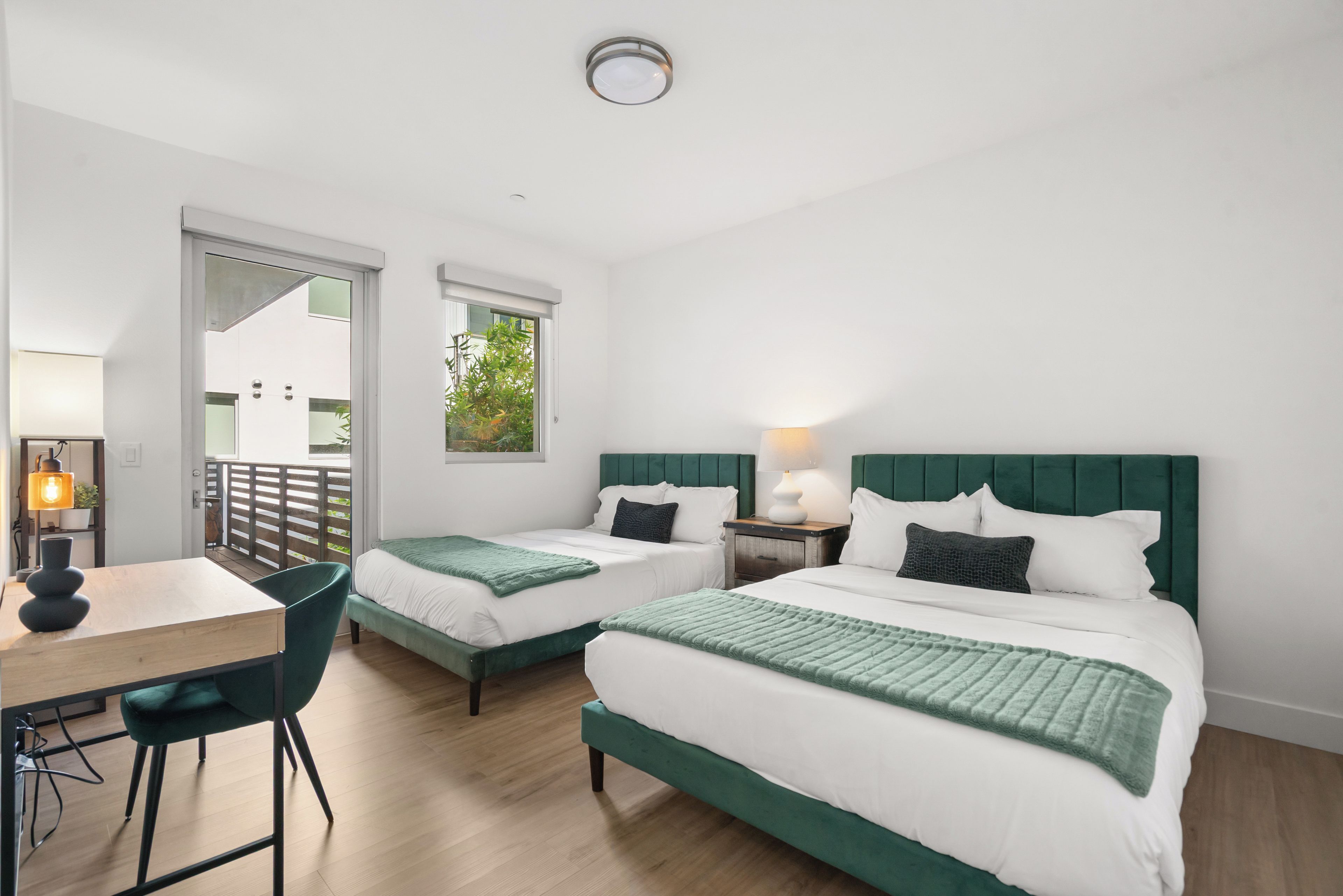 A hotel room with two beds, a desk, and a chair. There is a view of greenery outside the window.