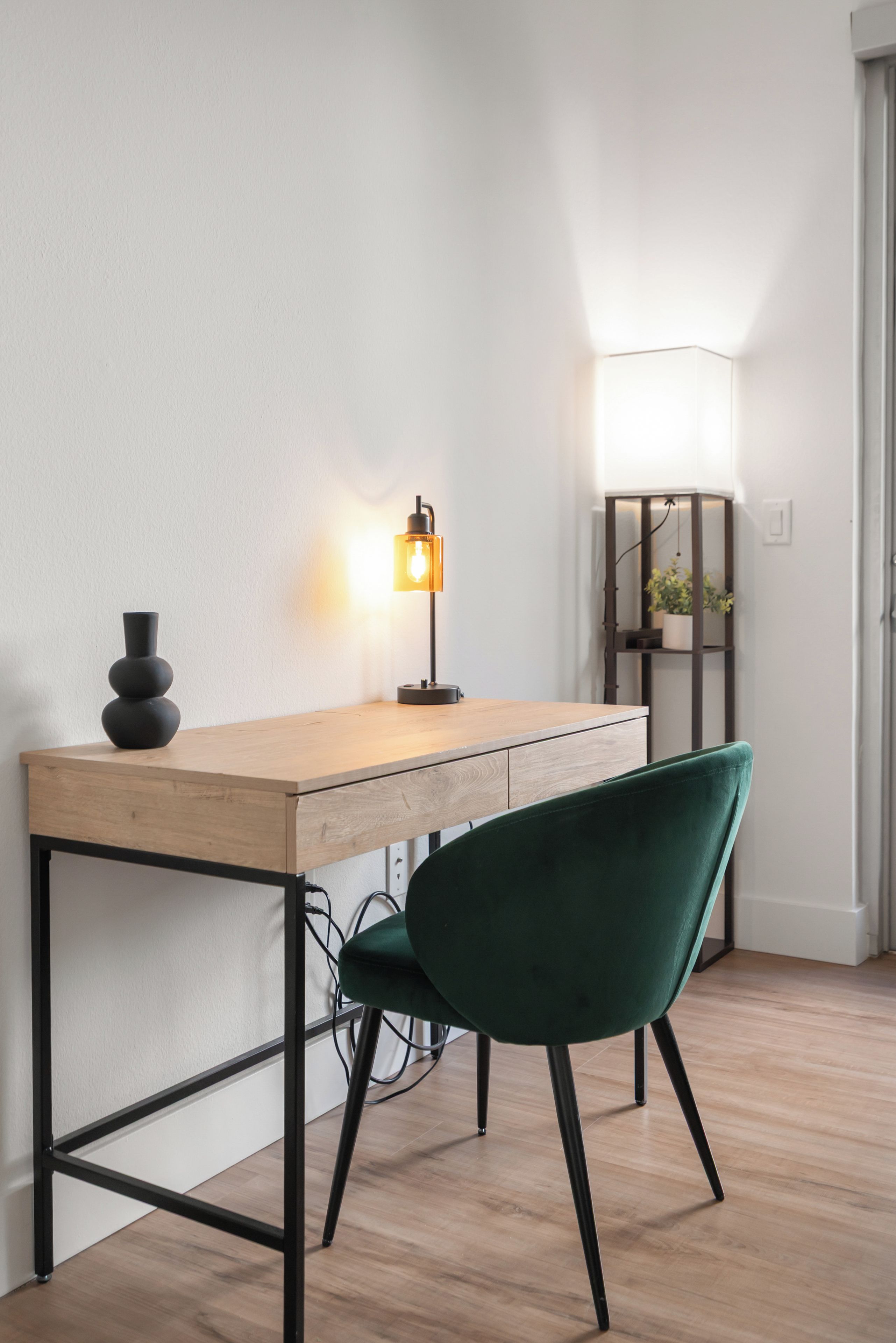 Ett modernt skrivbord med en träskiva och ett svart metallstativ, en grön sammetsstol, en svart vas och en golvlampa med en vit lampskärm.