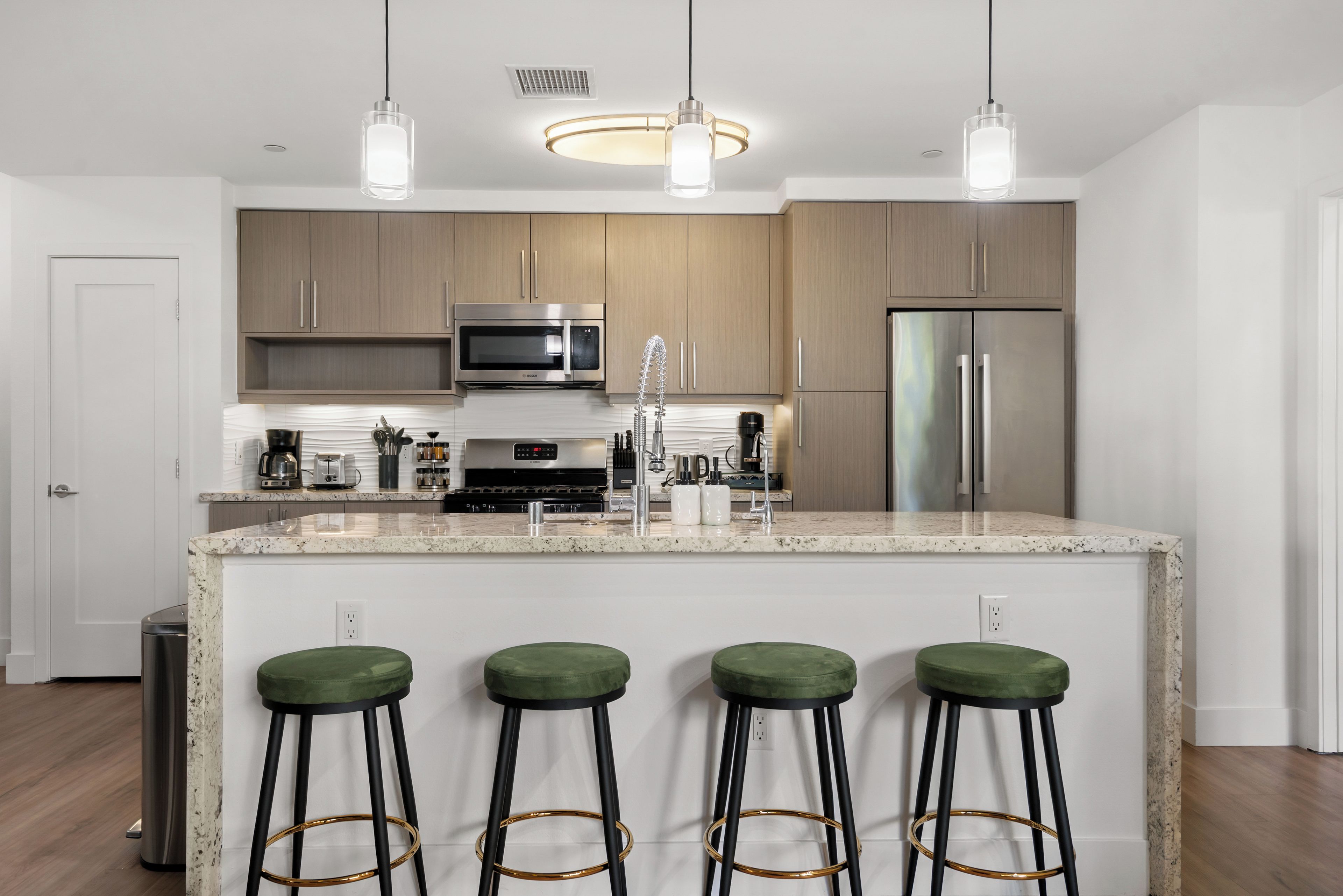 A modern kitchen with wooden cabinets, stainless steel appliances, a granite countertop, and green bar stools.