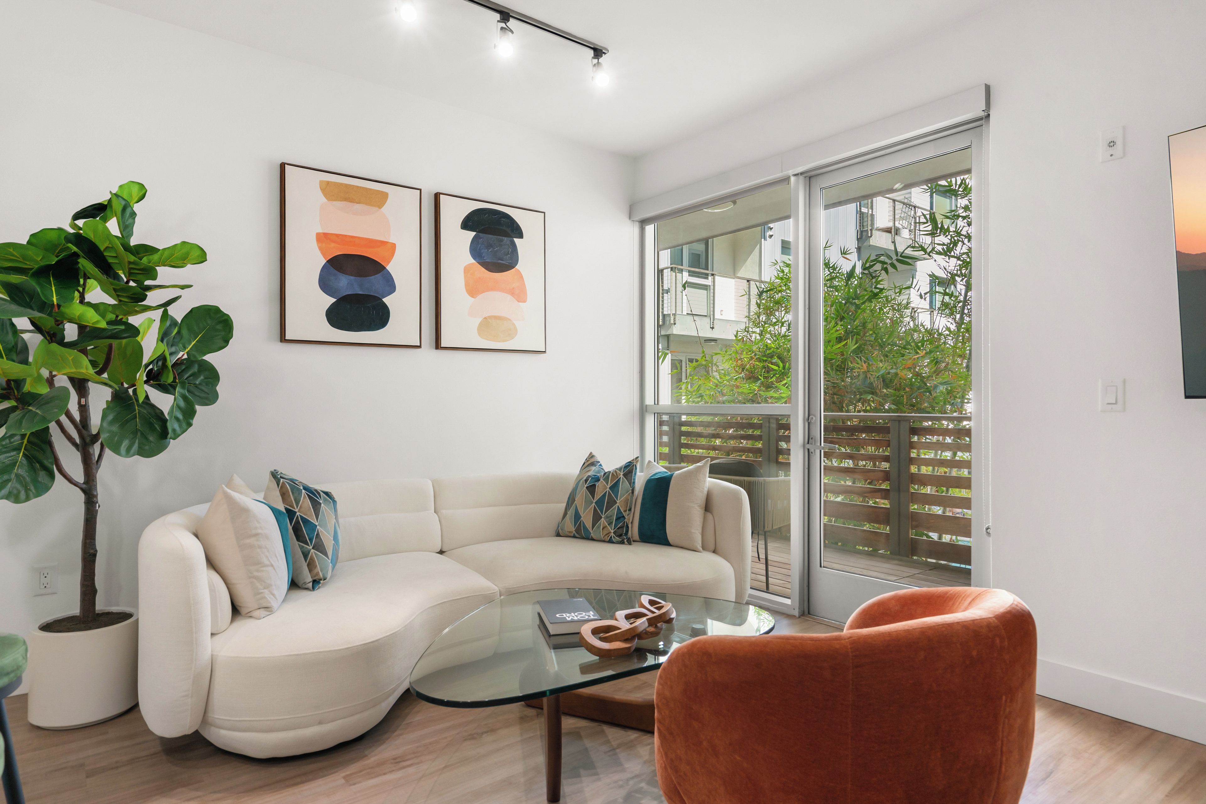 A modern living room with a curved sofa, a glass coffee table, and a large potted plant. There are abstract paintings on the wall and a sliding glass door leading to a balcony.