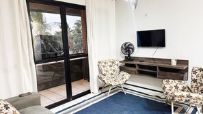 A living room with a sliding glass door, a flat-screen TV, a fan, and two patterned armchairs. - Comfort on Praia do Futuro by Reserva Mar (Fortaleza)