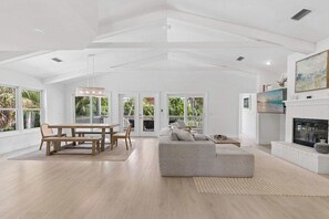 A spacious living room with a fireplace, a dining area with a wooden table and chairs, and large windows offering views of greenery. - Coastal Retreat - Sanibel Island Home, Pool and Beach Access! (Sanibel)