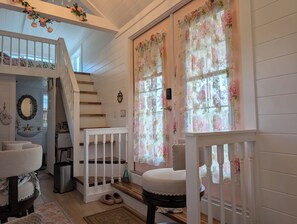 A staircase with a white railing leading to a landing, a small table with a chair, and a window with floral curtains. - Charming, Peaceful Tiny Home in Alachua, Florida (Alachua)